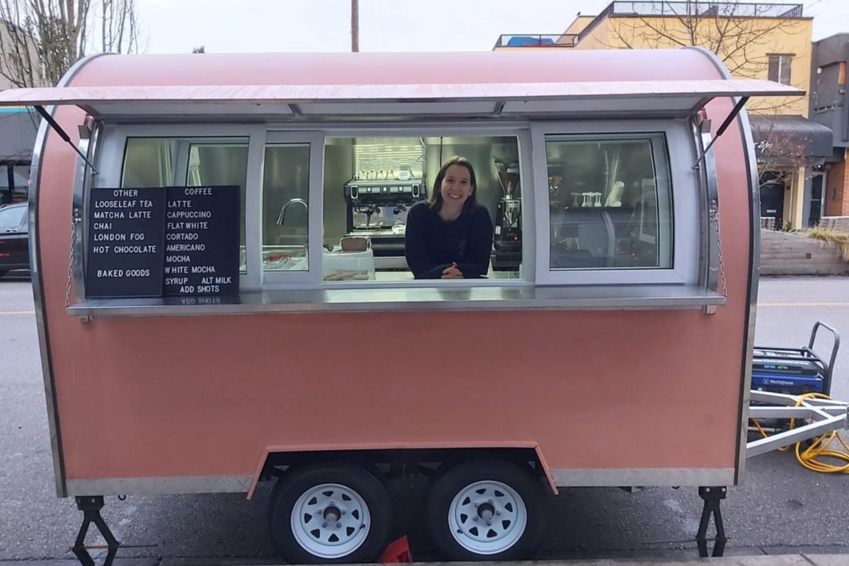 A salmon-pink coffee trailer parked on a city street, a barista leaning at the open service window with letterboard drinks menus mounted beside it