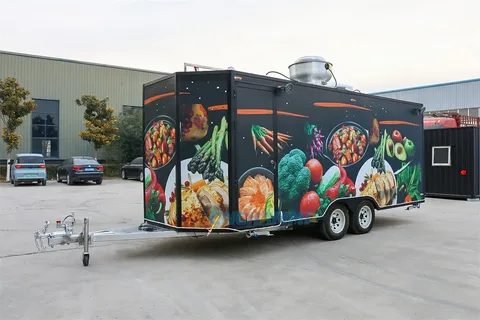 A black food trailer wrapped from end to end with photographs of prepared dishes and fresh vegetables, parked outside a workshop
