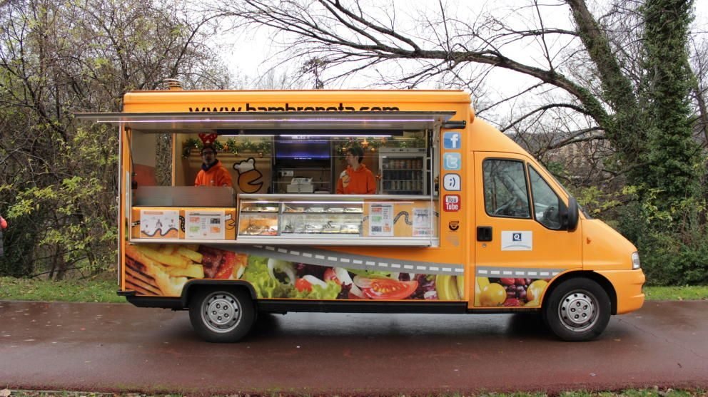 An orange truck-bodied mobile kitchen parked at the roadside with its side counter open and two staff serving from inside