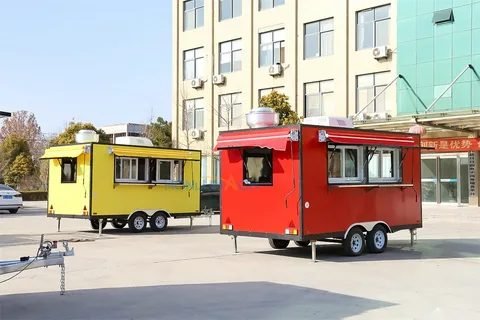 A yellow concession trailer and a red one parked side by side on a forecourt, both with tandem axles and awning-style serving hatches